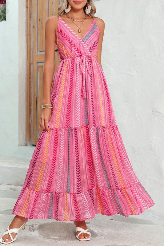 A woman wearing the Pink Tribal Tassel Tie V Neck Surplice Maxi Dress exudes an elegant vibe as she stands outdoors near a wooden door adorned with flowers above. Her white heels and matching handbag accentuate the bohemian style of her ensemble perfectly.