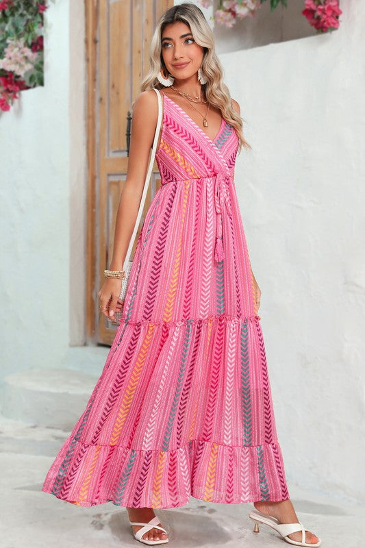 A woman wearing the Pink Tribal Tassel Tie V Neck Surplice Maxi Dress exudes an elegant vibe as she stands outdoors near a wooden door adorned with flowers above. Her white heels and matching handbag accentuate the bohemian style of her ensemble perfectly.