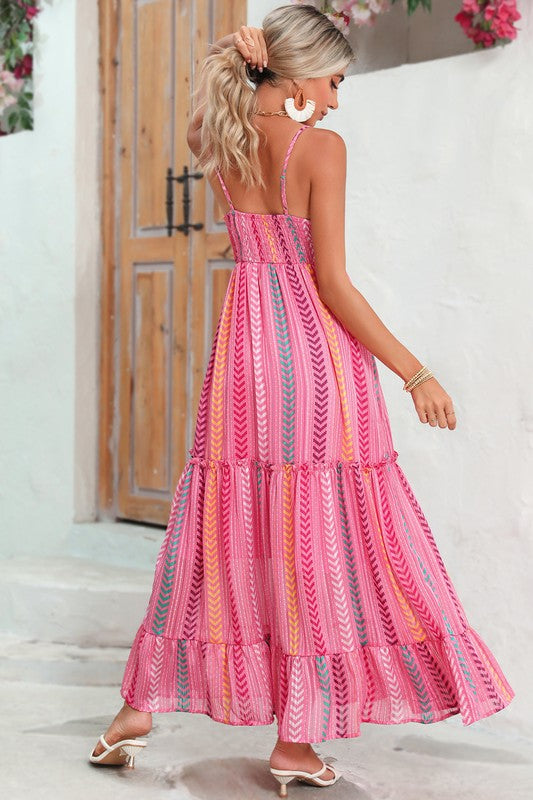 A woman wearing the Pink Tribal Tassel Tie V Neck Surplice Maxi Dress exudes an elegant vibe as she stands outdoors near a wooden door adorned with flowers above. Her white heels and matching handbag accentuate the bohemian style of her ensemble perfectly.