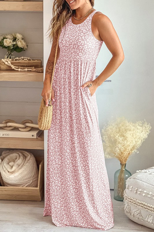 The Leopard Dust Baby Pink Sleeveless Maxi Dress, featuring a stylish white leopard print design on soft polyester fabric, elegantly drapes over a wooden hanger against a pristine white backdrop.