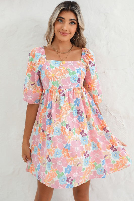 The Square Neck Bubble Puff Sleeve Floral Coral Dress, featuring a vibrant floral design on a light pink background, is elegantly displayed on a hanger against a plain white wall.