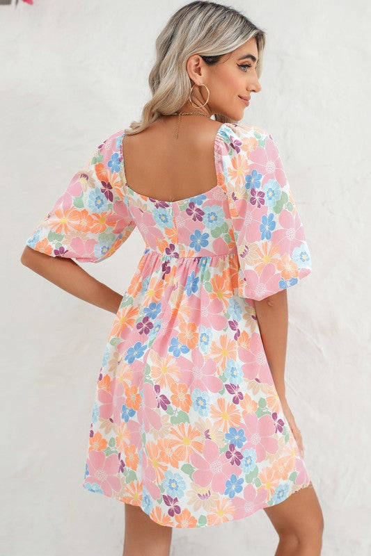 The Square Neck Bubble Puff Sleeve Floral Coral Dress, featuring a vibrant floral design on a light pink background, is elegantly displayed on a hanger against a plain white wall.