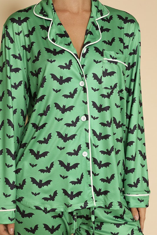 A person wearing the Halloween two-piece pajama set, featuring original prints of bats and a button-front long sleeve top in green, stands against a beige background, paired with black slippers decorated with pumpkin faces.