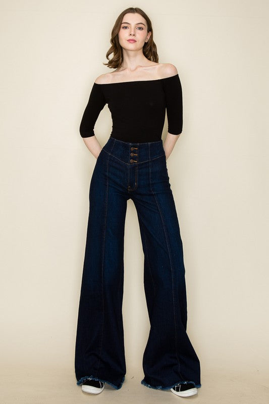 A person wearing high-waisted, Wide Leg Denim Pants in dark blue stretch denim fabric with front buttons and visible seams, paired with a black top and white sneakers, stands against a plain background.