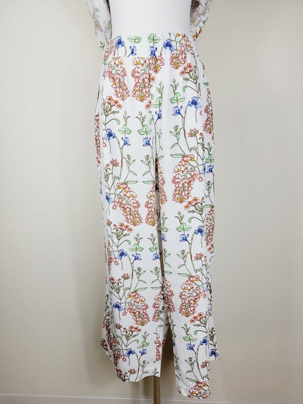 White pants with a vibrant floral pattern showcasing shades of blue, green, red, and pink. Made from airy cotton fabric, these wide-leg pants come with an elastic waistband for ultimate comfort.