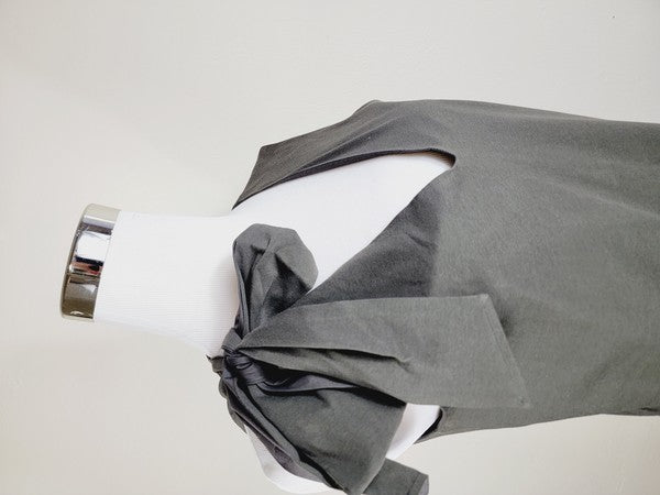 A Cotton Jersey dress in dark grey Turkish cotton, displayed on a mannequin, featuring an adjustable tie shoulder with a large bow on the left side and pockets, set against a plain white background.