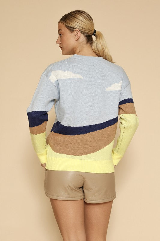 A woman stands against a beige background, wearing the Roadrunner knit sweater, featuring an exclusive in-house road runner intarsia design, and beige shorts.