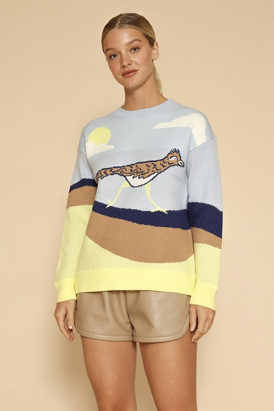 A woman stands against a beige background, wearing the Roadrunner knit sweater, featuring an exclusive in-house road runner intarsia design, and beige shorts.