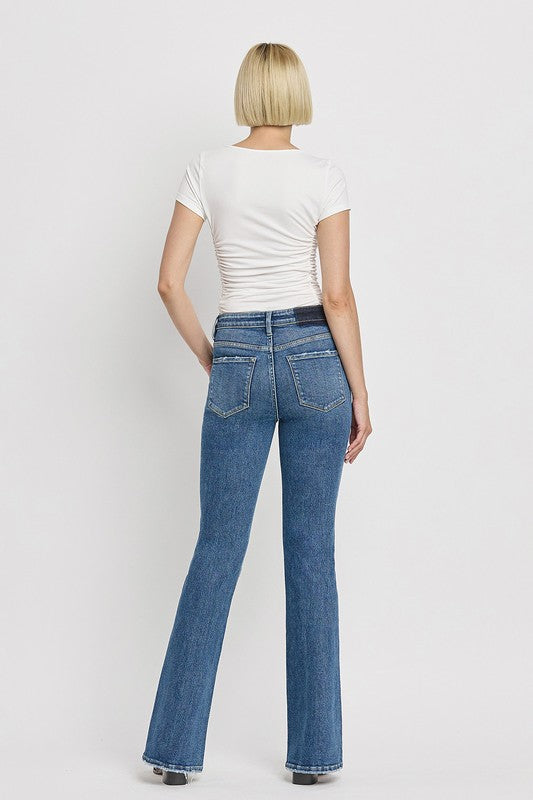 A person is wearing the High Rise Slant Hem Flare Jeans crafted from stretch denim, matched with black sandals, and standing against a plain white backdrop. The jeans' flattering fit enhances a chic and contemporary look.