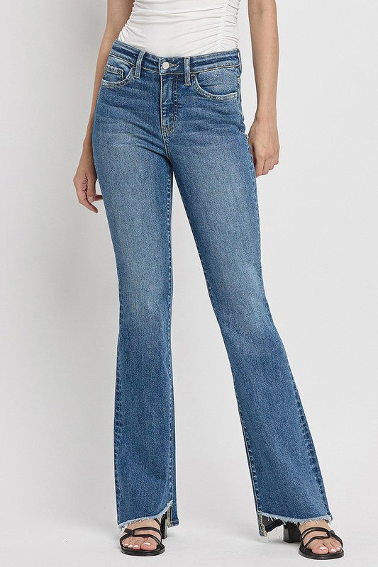 A person is wearing the High Rise Slant Hem Flare Jeans crafted from stretch denim, matched with black sandals, and standing against a plain white backdrop. The jeans' flattering fit enhances a chic and contemporary look.