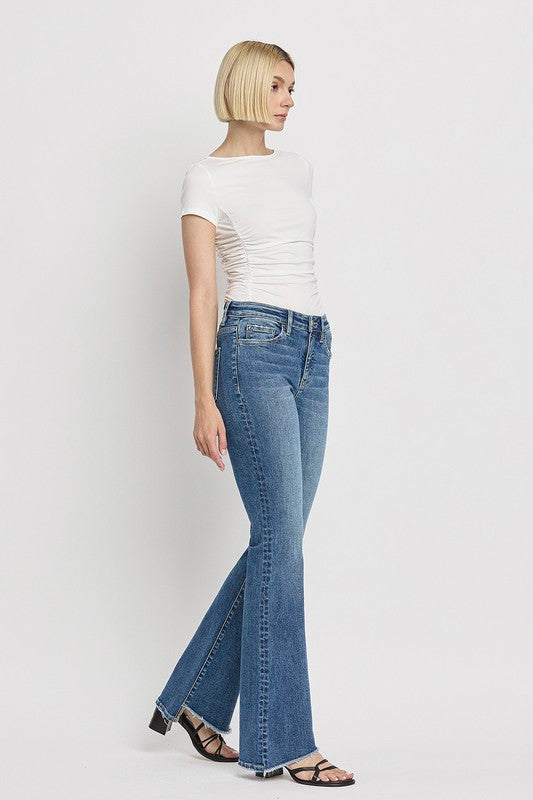 A person is wearing the High Rise Slant Hem Flare Jeans crafted from stretch denim, matched with black sandals, and standing against a plain white backdrop. The jeans' flattering fit enhances a chic and contemporary look.