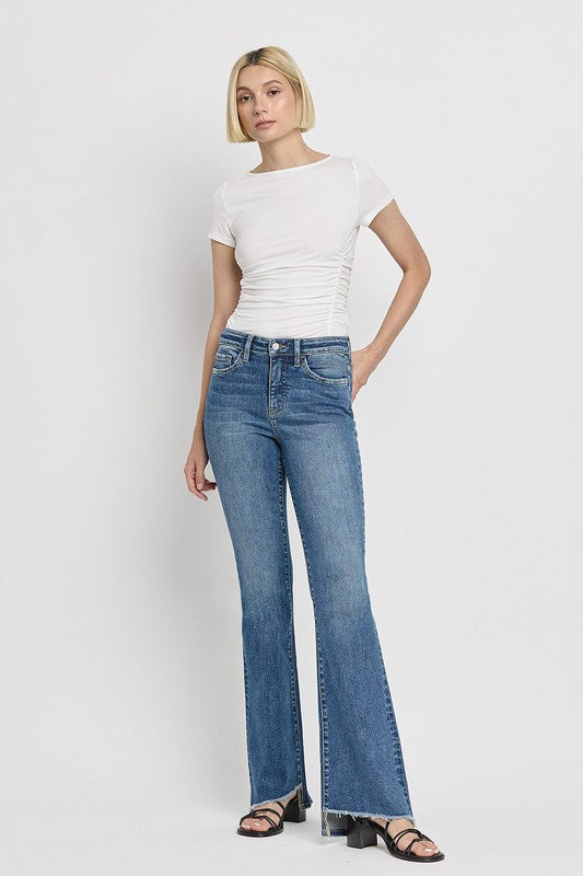 A person is wearing the High Rise Slant Hem Flare Jeans crafted from stretch denim, matched with black sandals, and standing against a plain white backdrop. The jeans' flattering fit enhances a chic and contemporary look.