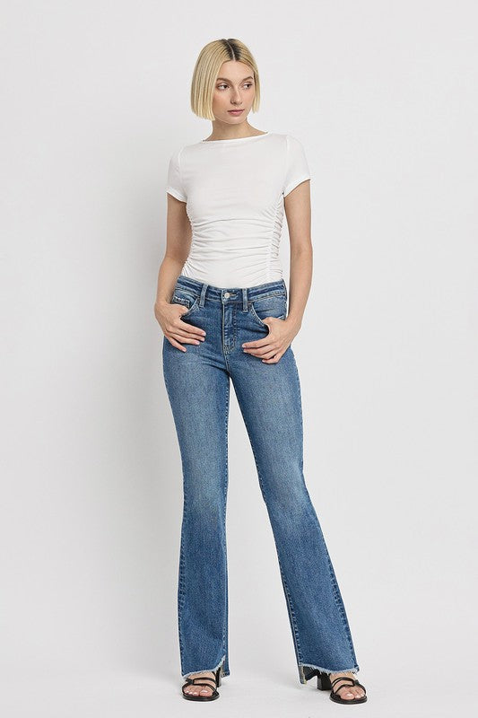 A person is wearing the High Rise Slant Hem Flare Jeans crafted from stretch denim, matched with black sandals, and standing against a plain white backdrop. The jeans' flattering fit enhances a chic and contemporary look.