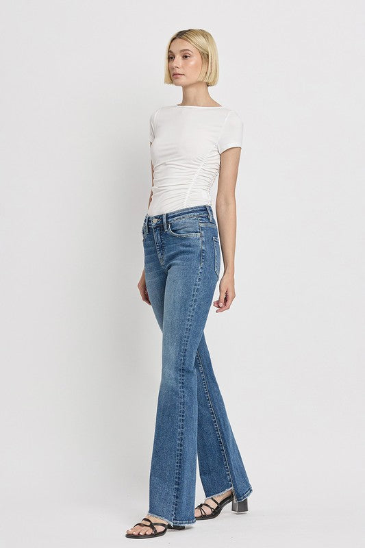 A person is wearing the High Rise Slant Hem Flare Jeans crafted from stretch denim, matched with black sandals, and standing against a plain white backdrop. The jeans' flattering fit enhances a chic and contemporary look.