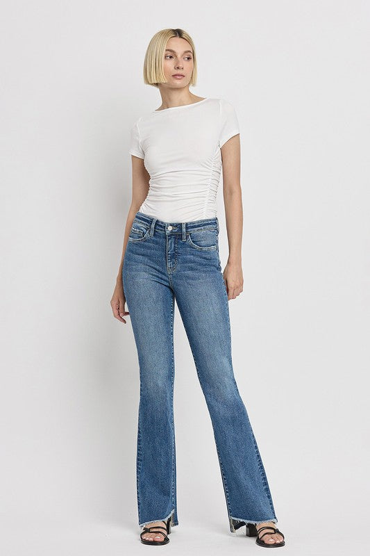 A person is wearing the High Rise Slant Hem Flare Jeans crafted from stretch denim, matched with black sandals, and standing against a plain white backdrop. The jeans' flattering fit enhances a chic and contemporary look.