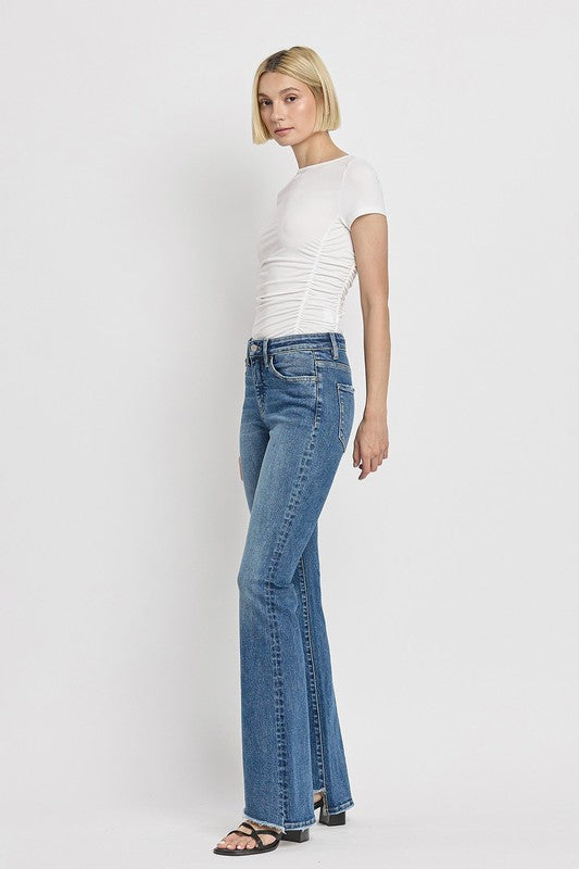 A person is wearing the High Rise Slant Hem Flare Jeans crafted from stretch denim, matched with black sandals, and standing against a plain white backdrop. The jeans' flattering fit enhances a chic and contemporary look.