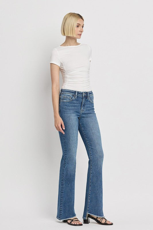 A person is wearing the High Rise Slant Hem Flare Jeans crafted from stretch denim, matched with black sandals, and standing against a plain white backdrop. The jeans' flattering fit enhances a chic and contemporary look.