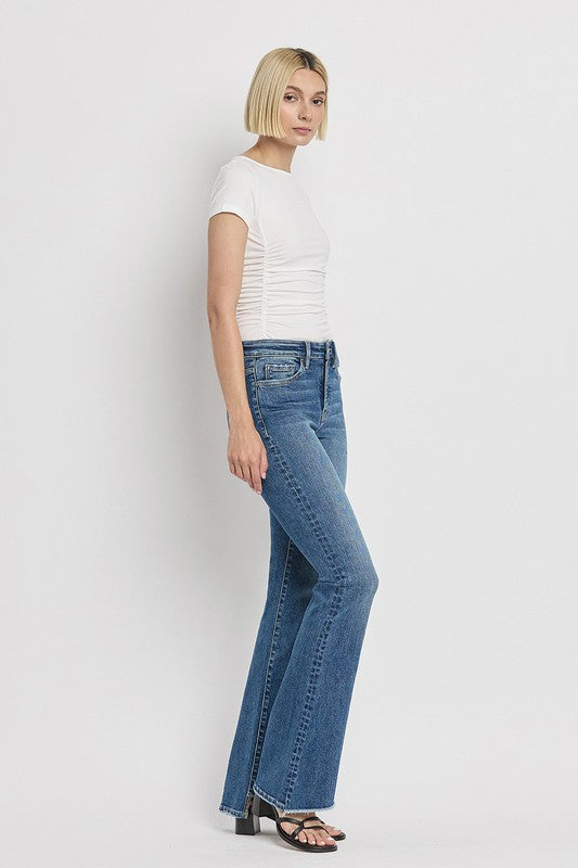 A person is wearing the High Rise Slant Hem Flare Jeans crafted from stretch denim, matched with black sandals, and standing against a plain white backdrop. The jeans' flattering fit enhances a chic and contemporary look.