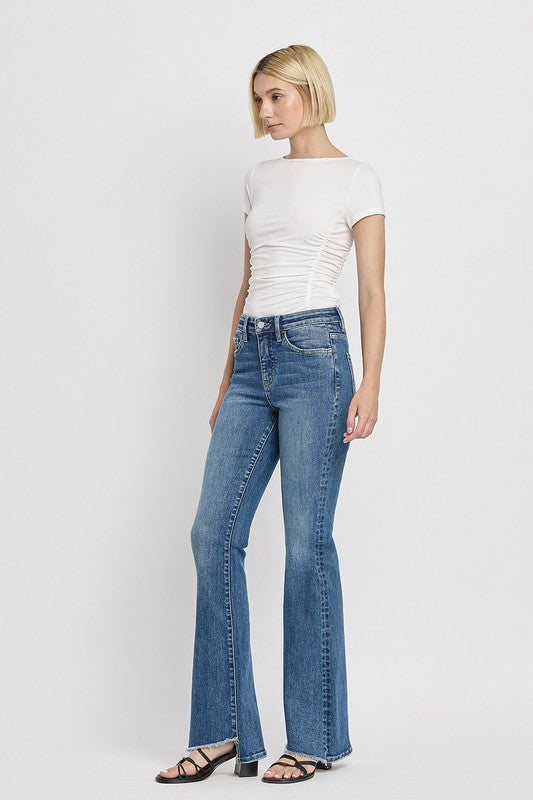 A person is wearing the High Rise Slant Hem Flare Jeans crafted from stretch denim, matched with black sandals, and standing against a plain white backdrop. The jeans' flattering fit enhances a chic and contemporary look.