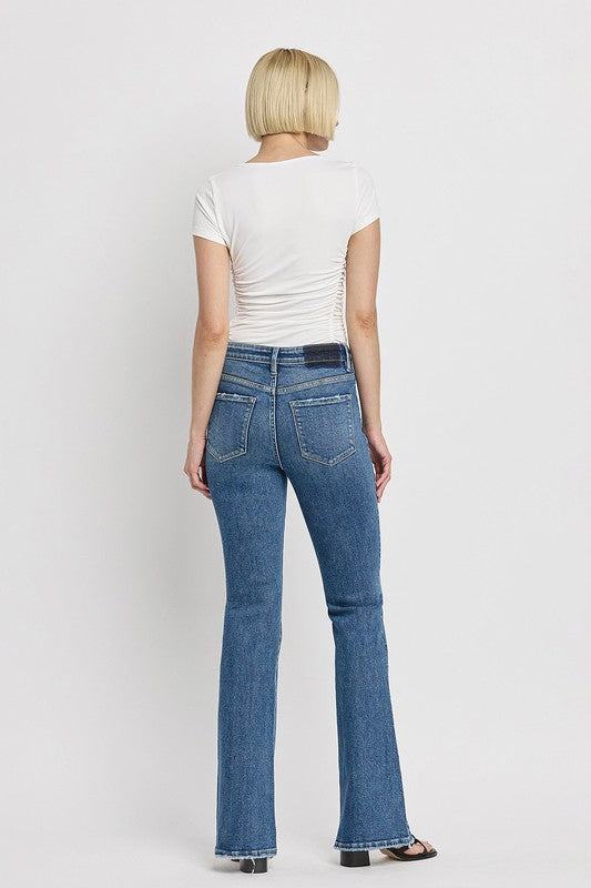 A person is wearing the High Rise Slant Hem Flare Jeans crafted from stretch denim, matched with black sandals, and standing against a plain white backdrop. The jeans' flattering fit enhances a chic and contemporary look.