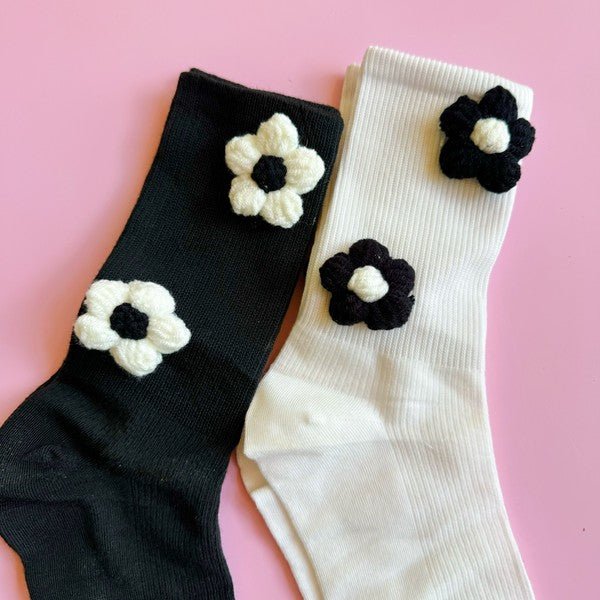 The Puffy Daisy Embellished Socks Set Of 2 includes two pairs of socks positioned on a pink surface: one black pair adorned with white knitted flowers and one white pair featuring black knitted flowers, highlighting a chic black and white contrast design.