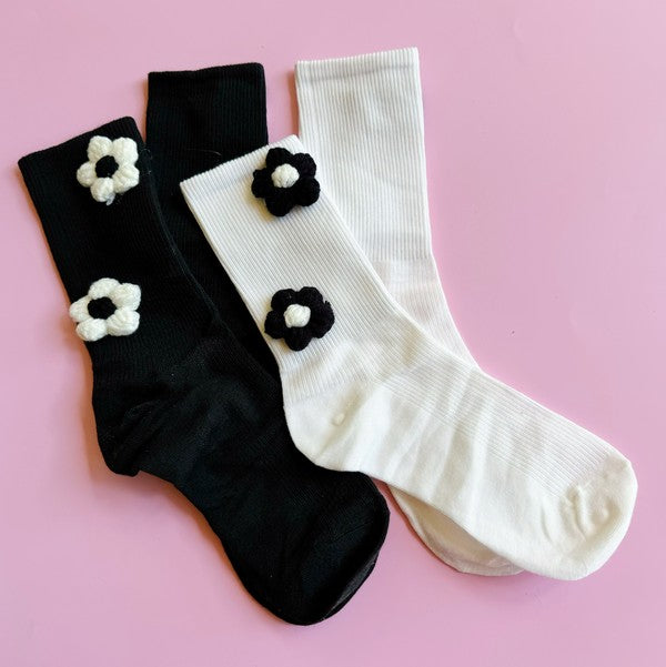 The Puffy Daisy Embellished Socks Set Of 2 includes two pairs of socks positioned on a pink surface: one black pair adorned with white knitted flowers and one white pair featuring black knitted flowers, highlighting a chic black and white contrast design.