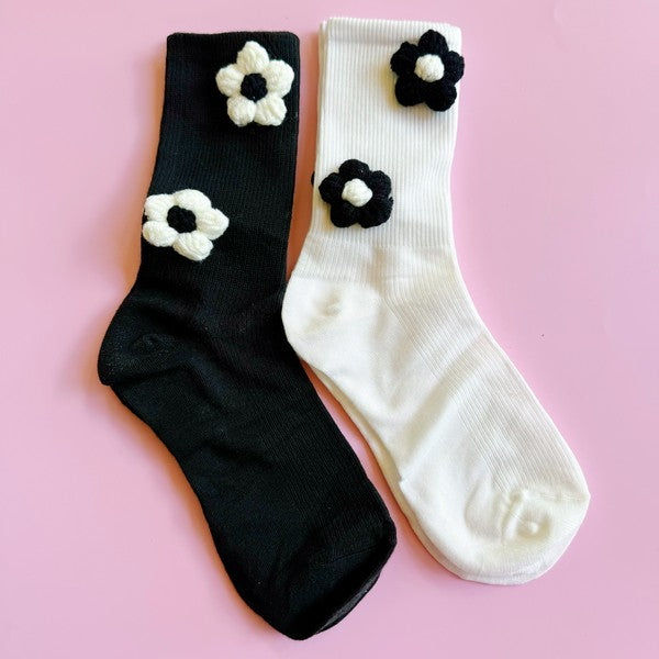 The Puffy Daisy Embellished Socks Set Of 2 includes two pairs of socks positioned on a pink surface: one black pair adorned with white knitted flowers and one white pair featuring black knitted flowers, highlighting a chic black and white contrast design.