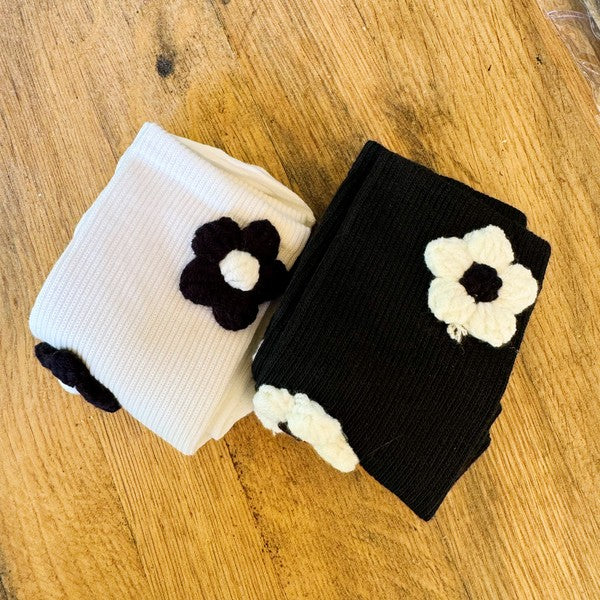 The Puffy Daisy Embellished Socks Set Of 2 includes two pairs of socks positioned on a pink surface: one black pair adorned with white knitted flowers and one white pair featuring black knitted flowers, highlighting a chic black and white contrast design.