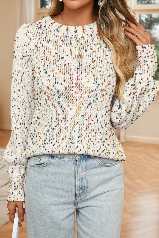 A person wearing a Space Dye Drop Shoulder Sweater, featuring a dotted print design in white and speckled knit, paired with light blue jeans stands indoors. Only the chest, arms, and midsection are visible. The room's background includes a wooden floor and part of a window.