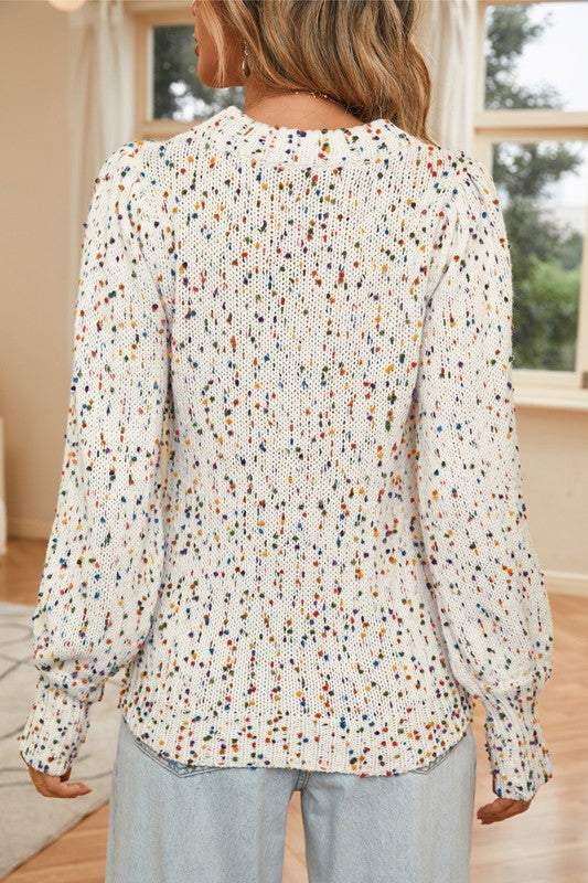 A person wearing a Space Dye Drop Shoulder Sweater, featuring a dotted print design in white and speckled knit, paired with light blue jeans stands indoors. Only the chest, arms, and midsection are visible. The room's background includes a wooden floor and part of a window.