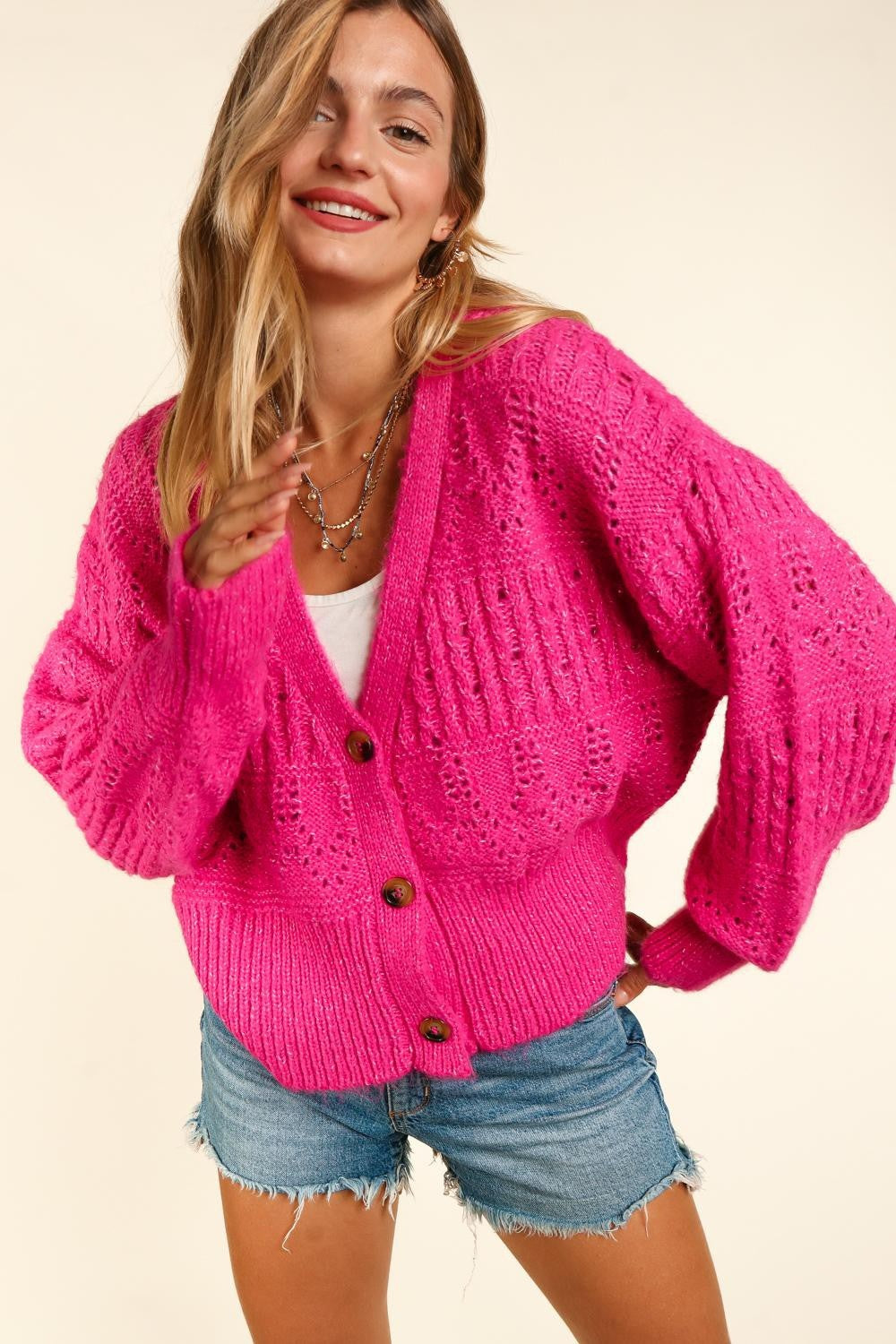 A person with long hair wears the trendy, bright pink Haptics Full Size Button Down Crop Long Sleeve Cardigan over a white top and denim shorts. They hold one side of the cardigan's collar and smile at the camera, showcasing their stylish and versatile layering.