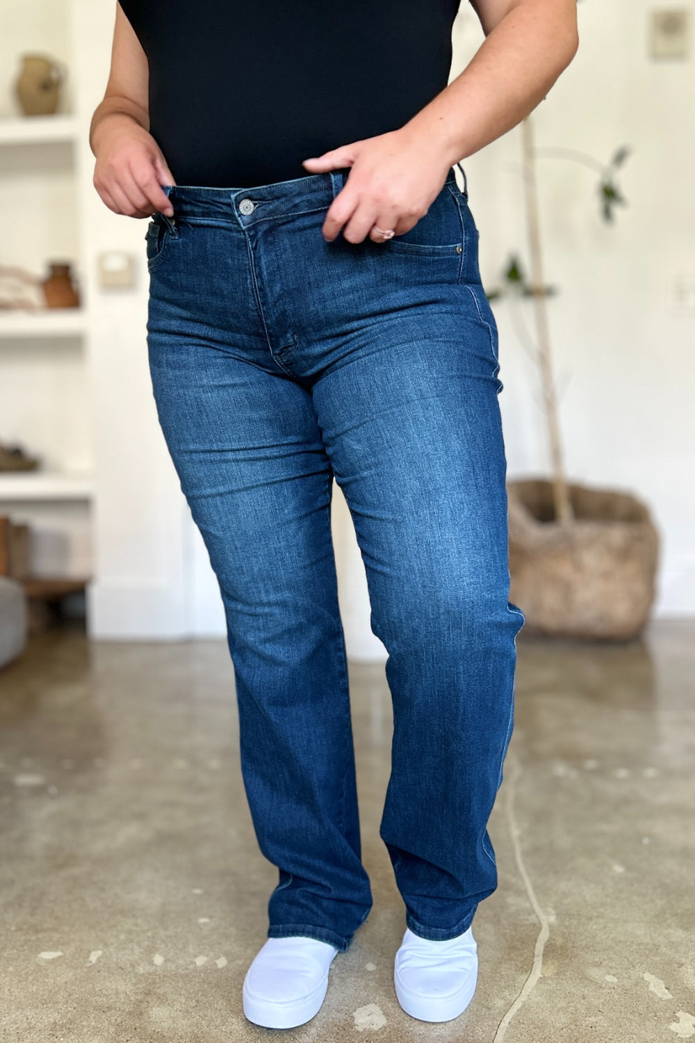 A person wearing a black top and Judy Blue Full Size Tummy Control Straight Jeans stands indoors with one hand on their hip. Their shoes are black with white soles. A plant and shelves are in the background.