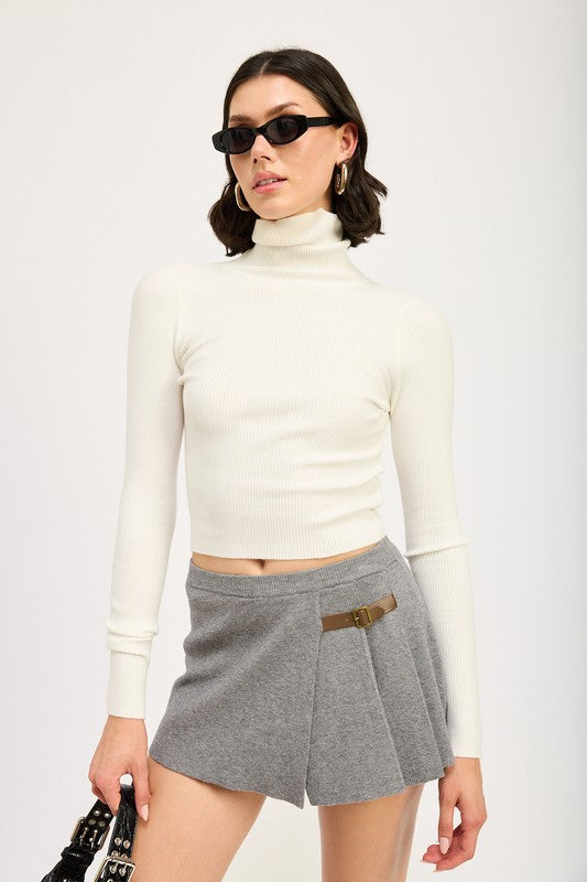 A person stands in a white turtleneck, PLEATED MINI SKORT, black loafers, and white socks. They accessorize with sunglasses and a small black handbag, adding a touch of sophistication to their size small ensemble.