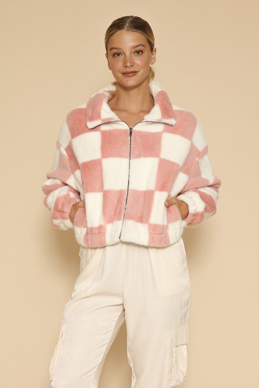 A woman standing against a beige background, showcasing an exclusive design: the Fuzzy checkered zip front jacket in pink and white, paired with light-colored pants.