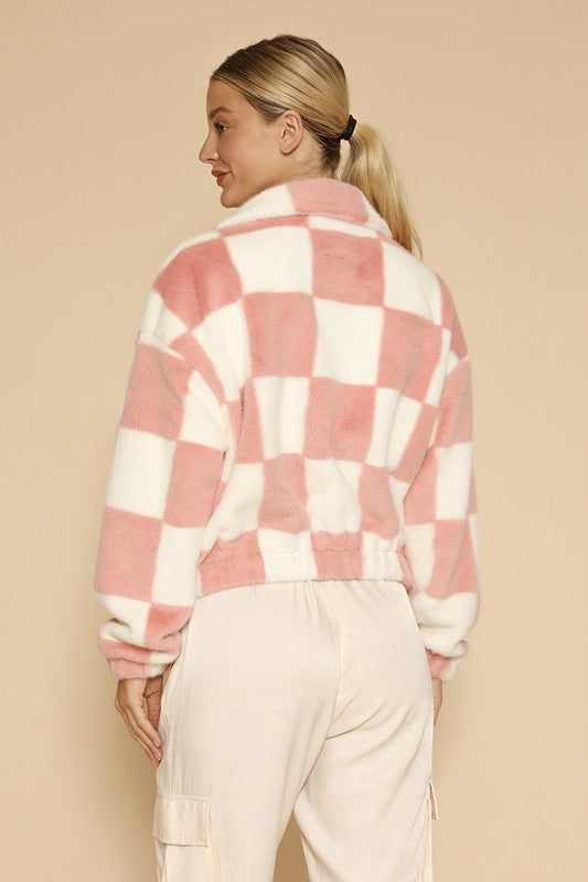 A woman standing against a beige background, showcasing an exclusive design: the Fuzzy checkered zip front jacket in pink and white, paired with light-colored pants.