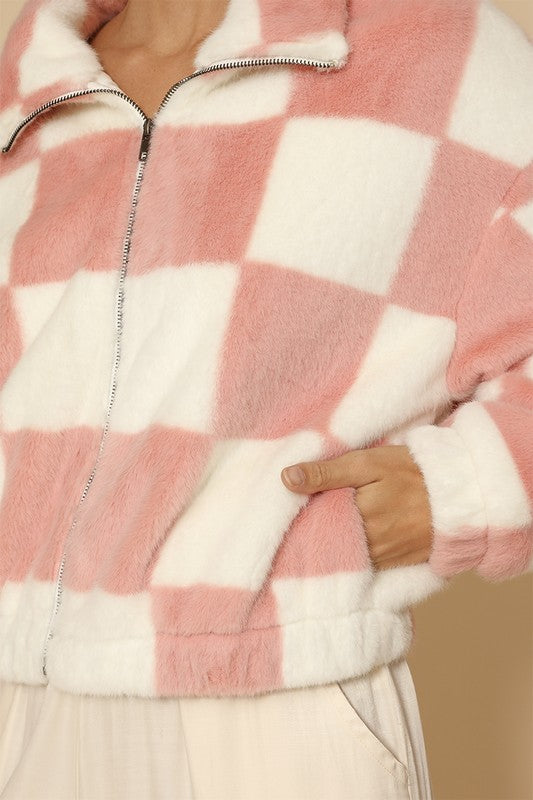 A woman standing against a beige background, showcasing an exclusive design: the Fuzzy checkered zip front jacket in pink and white, paired with light-colored pants.