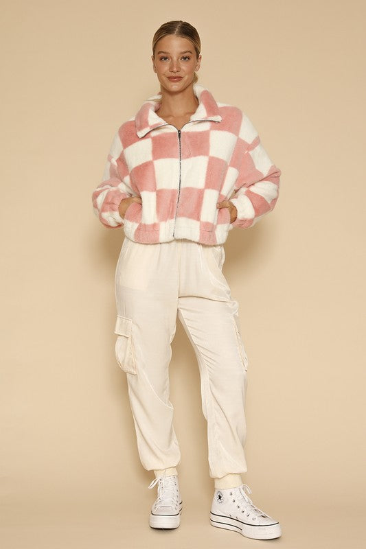 A woman standing against a beige background, showcasing an exclusive design: the Fuzzy checkered zip front jacket in pink and white, paired with light-colored pants.