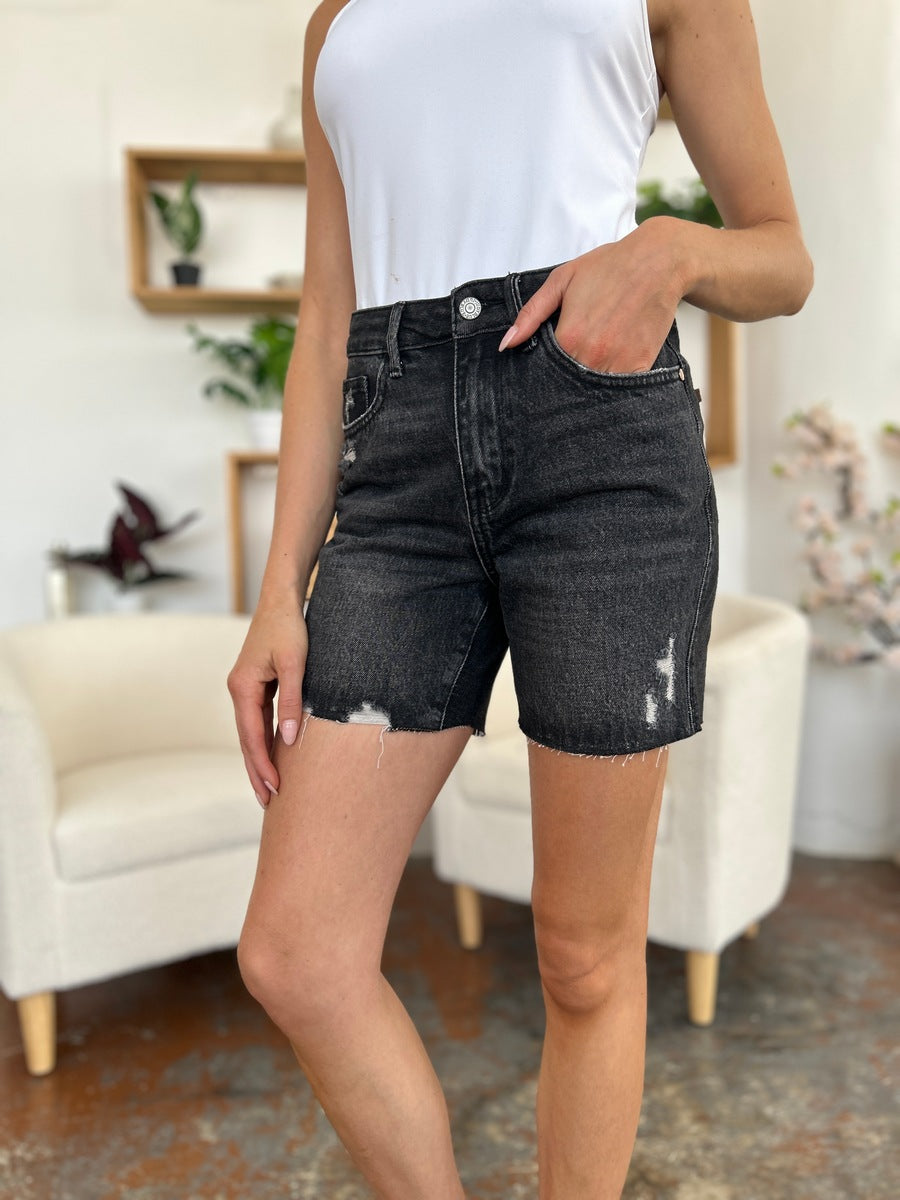 A person wearing a white t-shirt and Judy Blue Full Size High Waist Rigid Magic Denim Shorts is standing indoors in front of a beige chair.