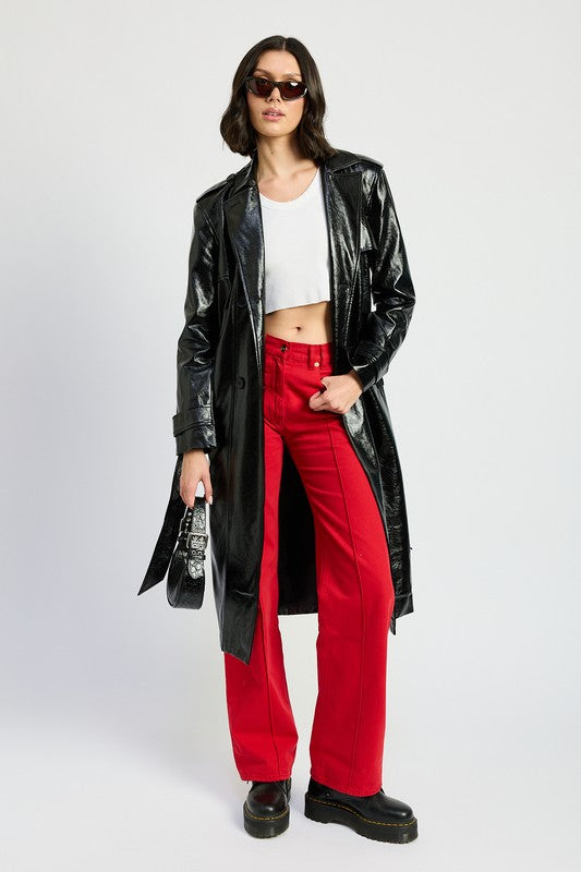 A woman is seen wearing a shiny FAUX LEATHER TRENCH COAT in black, paired with a white cropped top and red pants. She accessorizes her look with sunglasses and strikes a pose with one hand on her head. This stylish outfit is imported. The model is 5'9" tall and is wearing a size small.
