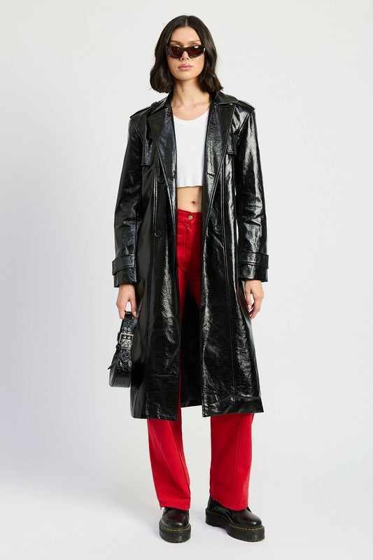 A woman is seen wearing a shiny FAUX LEATHER TRENCH COAT in black, paired with a white cropped top and red pants. She accessorizes her look with sunglasses and strikes a pose with one hand on her head. This stylish outfit is imported. The model is 5'9" tall and is wearing a size small.
