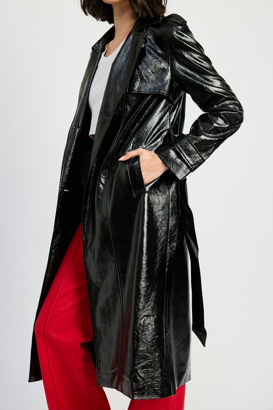 A woman is seen wearing a shiny FAUX LEATHER TRENCH COAT in black, paired with a white cropped top and red pants. She accessorizes her look with sunglasses and strikes a pose with one hand on her head. This stylish outfit is imported. The model is 5'9" tall and is wearing a size small.
