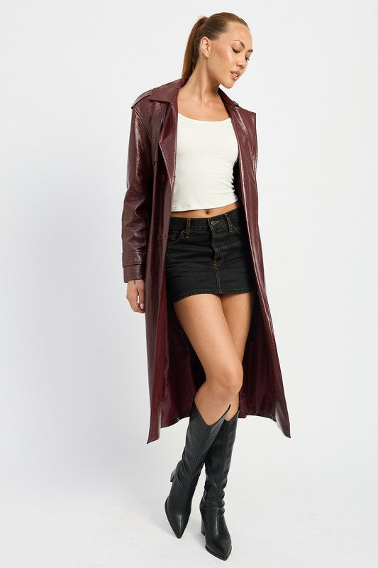 A woman is seen wearing a shiny FAUX LEATHER TRENCH COAT in black, paired with a white cropped top and red pants. She accessorizes her look with sunglasses and strikes a pose with one hand on her head. This stylish outfit is imported. The model is 5'9" tall and is wearing a size small.
