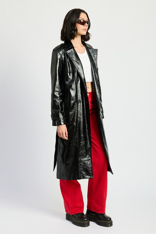 A woman is seen wearing a shiny FAUX LEATHER TRENCH COAT in black, paired with a white cropped top and red pants. She accessorizes her look with sunglasses and strikes a pose with one hand on her head. This stylish outfit is imported. The model is 5'9" tall and is wearing a size small.
