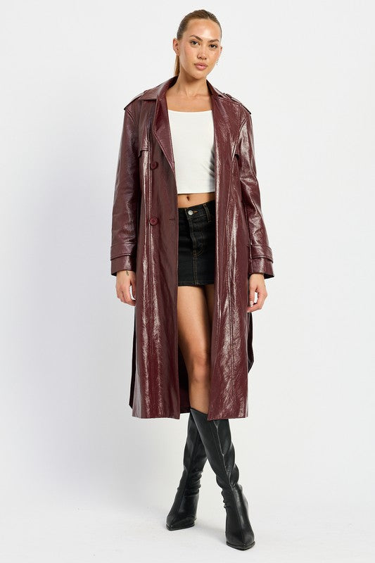 A woman is seen wearing a shiny FAUX LEATHER TRENCH COAT in black, paired with a white cropped top and red pants. She accessorizes her look with sunglasses and strikes a pose with one hand on her head. This stylish outfit is imported. The model is 5'9" tall and is wearing a size small.
