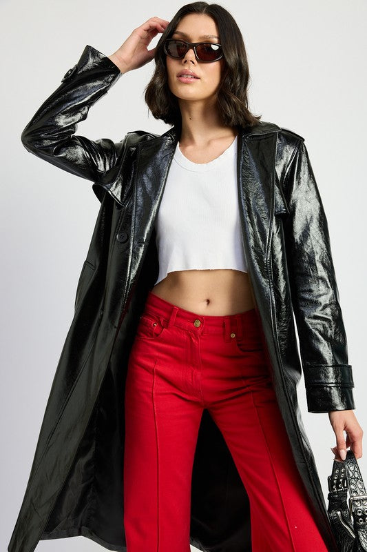 A woman is seen wearing a shiny FAUX LEATHER TRENCH COAT in black, paired with a white cropped top and red pants. She accessorizes her look with sunglasses and strikes a pose with one hand on her head. This stylish outfit is imported. The model is 5'9" tall and is wearing a size small.
