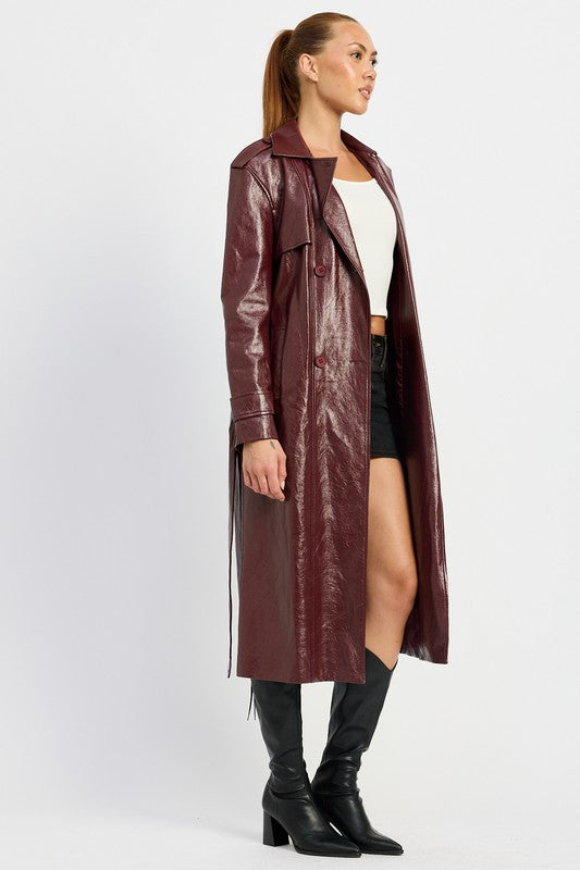 A woman is seen wearing a shiny FAUX LEATHER TRENCH COAT in black, paired with a white cropped top and red pants. She accessorizes her look with sunglasses and strikes a pose with one hand on her head. This stylish outfit is imported. The model is 5'9" tall and is wearing a size small.
