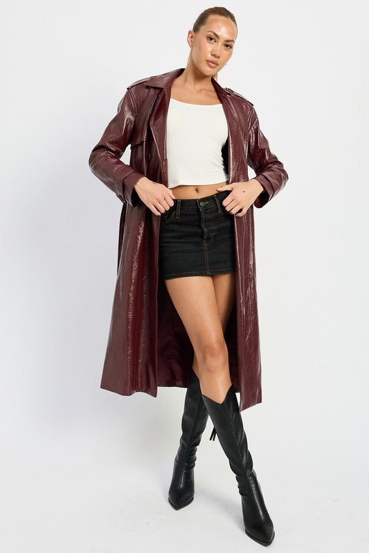 A woman is seen wearing a shiny FAUX LEATHER TRENCH COAT in black, paired with a white cropped top and red pants. She accessorizes her look with sunglasses and strikes a pose with one hand on her head. This stylish outfit is imported. The model is 5'9" tall and is wearing a size small.
