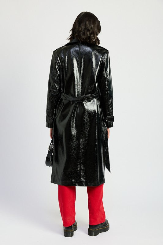 A woman is seen wearing a shiny FAUX LEATHER TRENCH COAT in black, paired with a white cropped top and red pants. She accessorizes her look with sunglasses and strikes a pose with one hand on her head. This stylish outfit is imported. The model is 5'9" tall and is wearing a size small.
