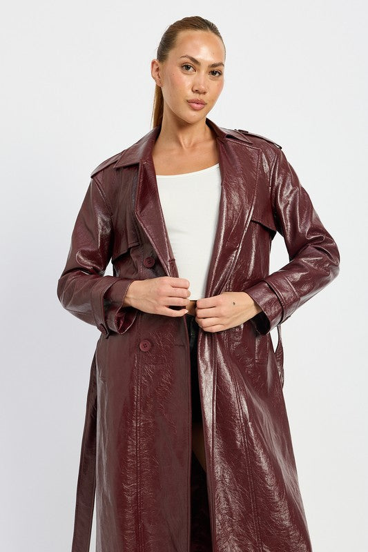 A woman is seen wearing a shiny FAUX LEATHER TRENCH COAT in black, paired with a white cropped top and red pants. She accessorizes her look with sunglasses and strikes a pose with one hand on her head. This stylish outfit is imported. The model is 5'9" tall and is wearing a size small.
