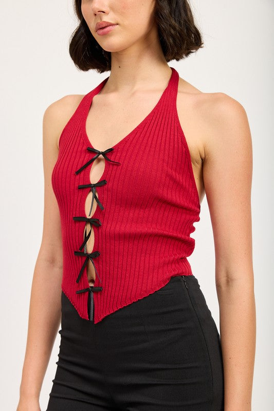 A woman wearing the HALTER NECK TOP WITH BOW DETAILS, a red halter top with black ribbon ties made from 70% viscose and 30% nylon, along with a black headband stands in front of a plain background.